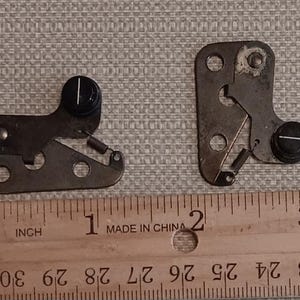 May include: Two metallic mechanical latch components with black circular knobs, each featuring a single white indicator line and a small tension spring, positioned above a wooden ruler showing measurements in inches up to three inches.