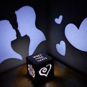 May include: A black wooden box with a light inside that projects a shadow of two silhouettes kissing and hearts on the wall. The box has the words "LOVE YOU LISA" engraved on it.