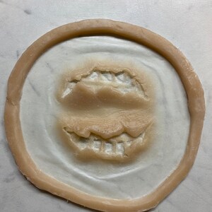 May include: A close-up of a beige silicone mold with two sets of teeth impressions. The mold is circular and has a raised edge.