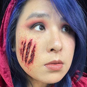 Peut inclure: Une femme aux cheveux bleus et une cape à capuche rouge a des marques de griffes réalistes sur la joue, avec des effets de sang et de déchirure de la peau.