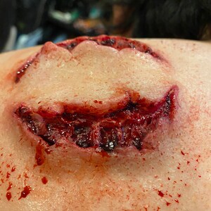 May include: A close-up of a fake, bloody wound on a person's shoulder. The wound is shaped like a mouth with teeth and is dripping blood.