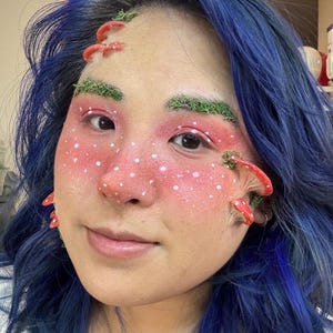 Könnte beinhalten: Eine Person mit blauen Haaren hat rotes und weißes Pilz-Make-up im Gesicht. Ihre Augenbrauen sind mit grünem Moos bedeckt.