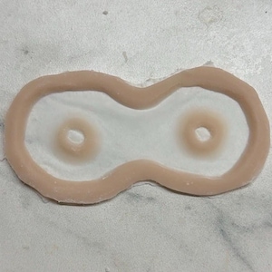 Puede incluir: Un molde de silicona con forma de ocho y dos hendiduras circulares en el centro. El molde tiene un borde exterior de color beige claro y una superficie interior blanca translúcida, diseñado para crear proyectos de manualidades detallados.