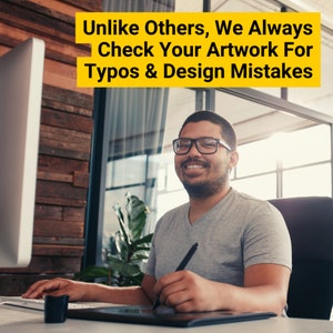 May include: A man wearing glasses smiles while using a digital drawing tablet and a stylus. He is sitting at a desk in front of a computer monitor. The text on the monitor reads "Unlike Others, We Always Check Your Artwork For Typos & Design Mistakes."
