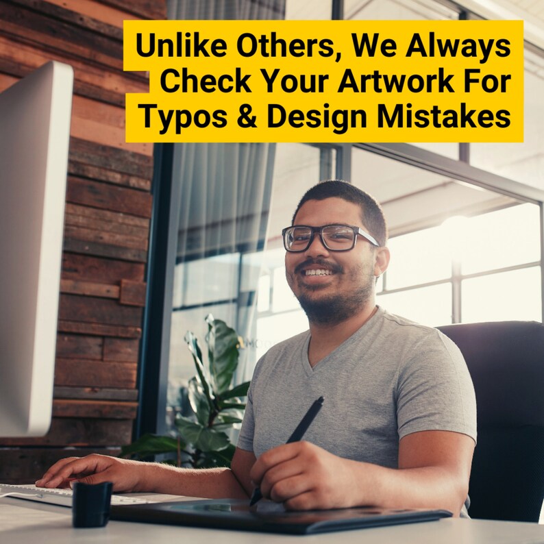 May include: A man wearing glasses smiles while using a digital drawing tablet and a stylus. He is sitting at a desk in front of a computer monitor. The text on the monitor reads "Unlike Others, We Always Check Your Artwork For Typos & Design Mistakes."