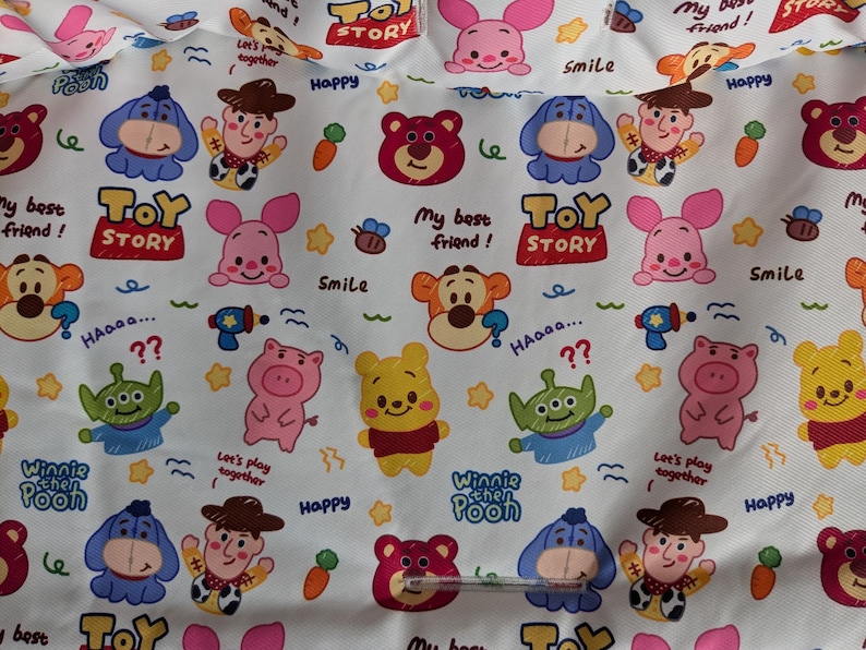 Puede incluir: Tela blanca con un patr&oacute;n repetitivo de personajes de dibujos animados de las pel&iacute;culas de Disney y Pixar. Los personajes incluyen Winnie the Pooh, Piglet, Tigger, Eeyore, personajes de Toy Story, y m&aacute;s. La tela est&aacute; impresa con el texto "Toy Story", "Winnie the Pooh", "&iexcl;Mi mejor amigo!" y "&iexcl;Juguemos juntos!".