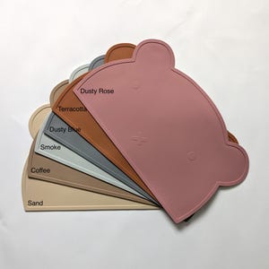 Könnte beinhalten: Ein Set aus sieben Silikon-Platzsets in verschiedenen Farben, darunter Dusty Rose, Terrakotta, Dusty Blue, Smoke, Coffee und Sand. Die Platzsets sind in Form eines Bären mit abgerundeten Ohren und einem kleinen Loch in der Mitte. Die Platzsets sind fächerförmig angeordnet, wobei das Dusty Rose-Platzset oben liegt.