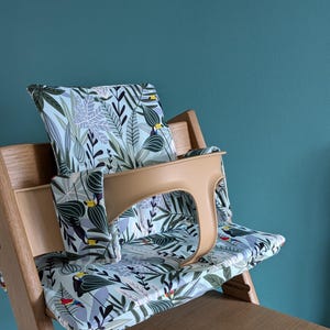 May include: A high chair with a light blue and green tropical leaf patterned cushion and a tan tray. The cushion has a toucan bird design.