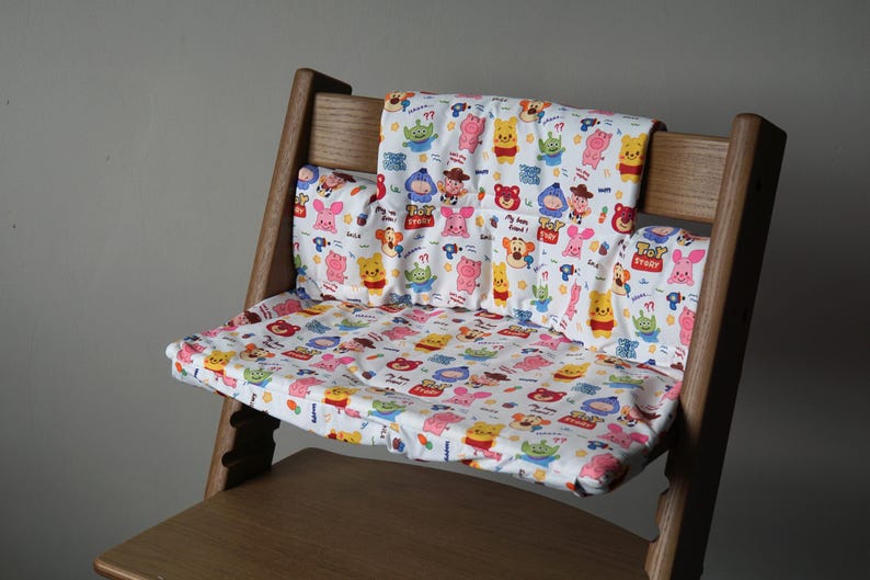 Puede incluir: Una silla alta de madera con una funda de asiento de tela blanca y colorida con personajes de dibujos animados de la franquicia Toy Story. La funda tiene una variedad de personajes, incluidos Woody, Buzz Lightyear y Jessie.