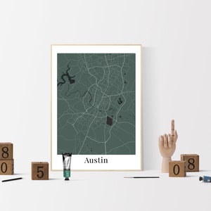 May include: A framed print of a map of Austin, Texas, with a green background and white lines. The print is on a white surface with wooden blocks spelling out the number 805 and a wooden hand sculpture. There is a tube of paint and a paintbrush on the surface.