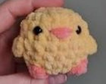 May include: A small, crocheted yellow chick with pink feet and a pink beak. The chick has black eyes and a fluffy texture.