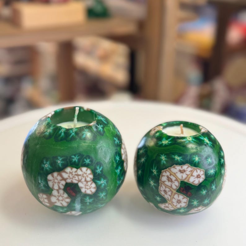 May include: Two green and white patterned sphere candles with a white wax centre. The candles are on a white surface.