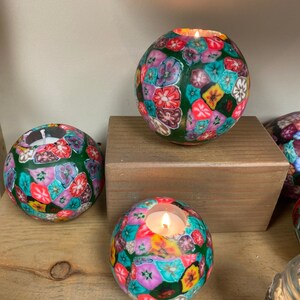 May include: Three round tealight candle holders with a green background and colorful floral pattern. The candle holders are made of a textured material and have a small opening at the top for the tealight candle.