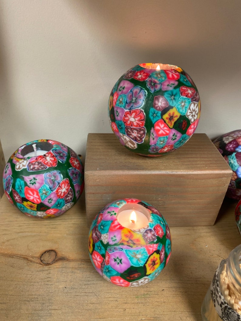 May include: Three round tealight candle holders with a green background and colorful floral pattern. The candle holders are made of a textured material and have a small opening at the top for the tealight candle.