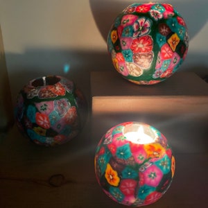 May include: Three round tealight candle holders with a colorful floral pattern. The candle holders are made of a textured material and have a green base.