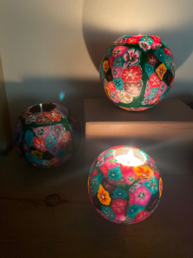 May include: Three round tealight candle holders with a colorful floral pattern. The candle holders are made of a textured material and have a green base.