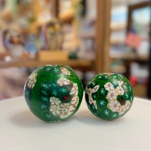 May include: Two green glass beads with white and gold floral patterns. The larger bead is in the foreground and the smaller bead is in the background.