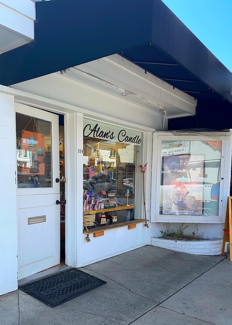 May include: A white shop front with a blue awning. The shop front has a glass door and two large windows. The windows display various items, including candles, a sign that says "Alan's Candle", and a sign that says "Alan's Candle Handmade".