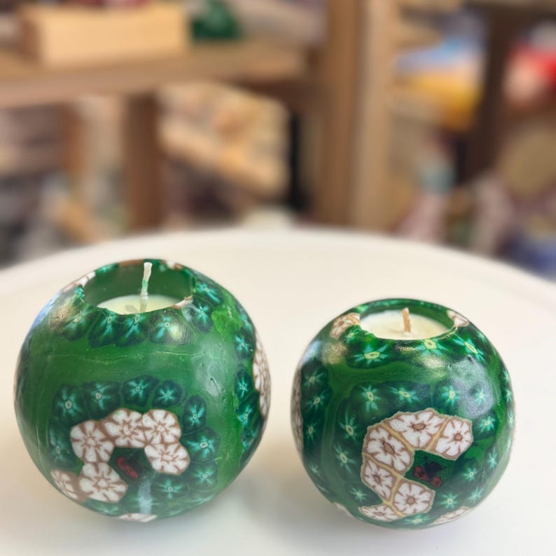 May include: Two green sphere candles with white and brown floral designs. The candles are sitting on a white surface.