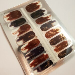 May include: A sheet of nail polish wraps with a gold, brown, and purple abstract design. The wraps are packaged in a silver foil pouch.