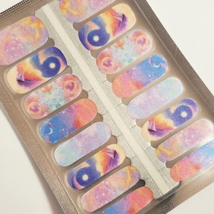 May include: A pack of nail polish wraps with a galaxy theme. The wraps feature a pink, blue, and purple galaxy design with white stars and a crescent moon.