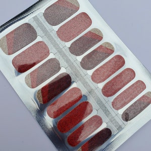 May include: A package of nail wraps in shades of red, brown, and gold. The nail wraps are rectangular with rounded edges and a glittery finish. The package is silver and has a clear window showing the nail wraps.