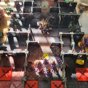 May include: A tabletop game board with a variety of miniature figures, including green, purple, and yellow figures. The board is made of a dark gray material and has a grid pattern. There are also several red squares with a white cross on them.