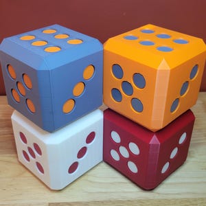 May include: Four large, colorful dice stacked on a wooden surface. The dice are blue, orange, white, and red, each with contrasting colored dots. The dice are a fun, geometric design.