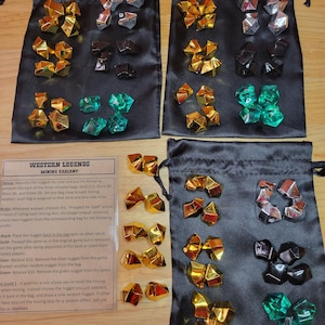 Puede incluir: Cuatro bolsas de terciopelo negro con pepitas de gemas doradas, plateadas, negras y verdes en su interior. Las bolsas están etiquetadas con 'Western Legends Mining Variant' y las reglas del juego impresas en una tarjeta.