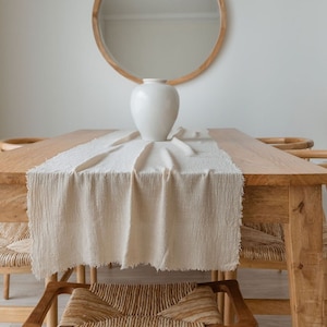 Peut inclure: Un vase en céramique blanche posé sur une table en bois avec un chemin de table en lin blanc. Un miroir rond encadré de bois est accroché au mur derrière le vase.