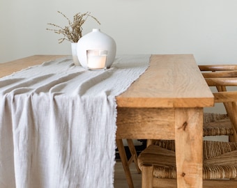 May include: A wooden dining table with a light gray linen table runner and two woven chairs. A white ceramic vase with a candle sits on the table runner.