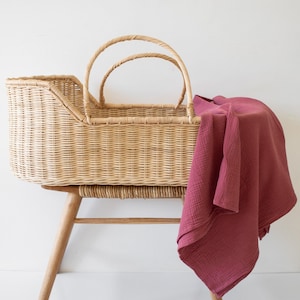 May include: A wicker bassinet with arched handles on a wooden stand. A burgundy muslin blanket is draped over the side. The bassinet and blanket are set against a white background.