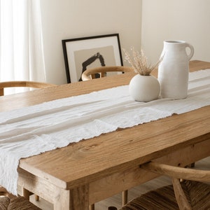 May include: A wooden dining table with a white linen table runner. Two chairs with woven seats are visible. A white vase with dried flowers and a white pitcher are on the table.