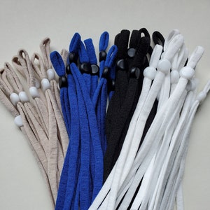 May include: A collection of adjustable face mask lanyards in various colors: beige, blue, black, and white. Each lanyard features a cord with a plastic adjuster and a loop for attaching to a mask. The lanyards are designed to keep masks accessible.