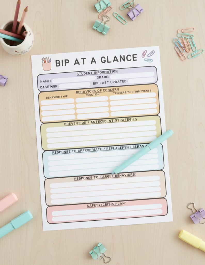 May include: A printed "BIP AT A GLANCE" form with sections for student information, behaviors, and safety plans. A light blue pen rests on the form, surrounded by colorful paper clips and markers. The form is on a light wood surface.