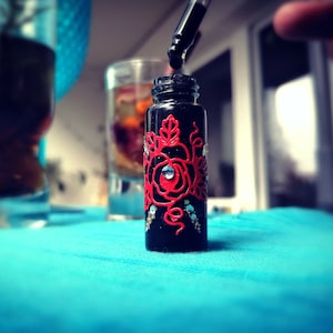 May include: A small black glass bottle with a red rose design and silver glitter accents. The bottle has a dropper attached to the top.