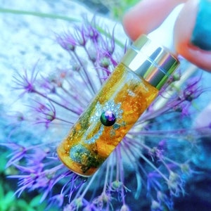 Op de afbeelding: Een klein, doorzichtig glazen flesje met een gouden dop en een druppelaar. Het flesje is gevuld met een gouden vloeistof en bevat glitter en een kleine, iriserende edelsteen. Het flesje ligt op een paarse bloem.