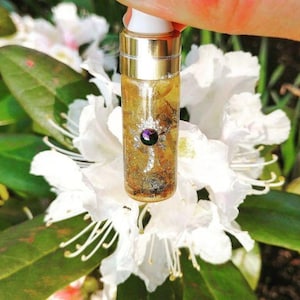 May include: A small glass bottle with a gold cap and a purple gemstone embedded in the front. The bottle is filled with a clear liquid and gold glitter. The bottle is held in front of a white flower.