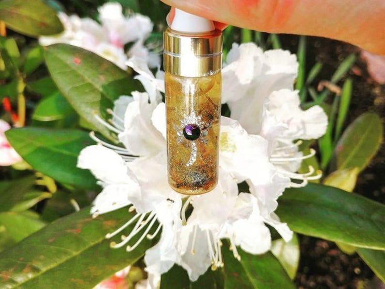 May include: A small glass bottle with a gold cap and a purple gemstone embedded in the front. The bottle is filled with a clear liquid and gold glitter. The bottle is held in front of a white flower.