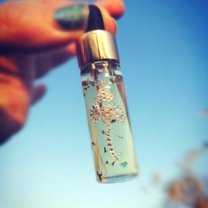 May include: A small, clear glass bottle with a gold cap and black dropper. The bottle contains a clear liquid with decorative gold and iridescent accents. The background is a bright blue sky.