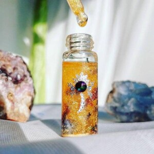 May include: A small glass bottle with a gold glitter and clear liquid inside. The bottle has a silver and black decorative design with a large iridescent gemstone in the center. The bottle is sitting on a white surface.