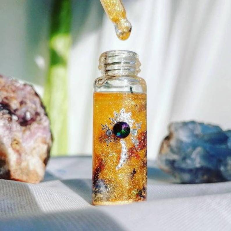 May include: A small glass bottle with a gold glitter and clear liquid inside. The bottle has a silver and black decorative design with a large iridescent gemstone in the center. The bottle is sitting on a white surface.