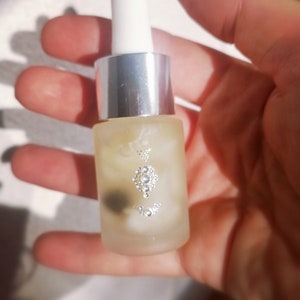 May include: A clear glass bottle with a silver cap and a white dropper. The bottle contains a light yellow liquid and has a small, silver, decorative design on the front.