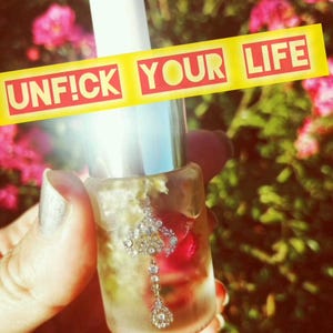May include: A clear glass bottle with a silver cap and a white dropper. The bottle contains a clear liquid with small white flowers and a red rose petal. The bottle is labeled "Unf!ck Your Life" in yellow and red letters.