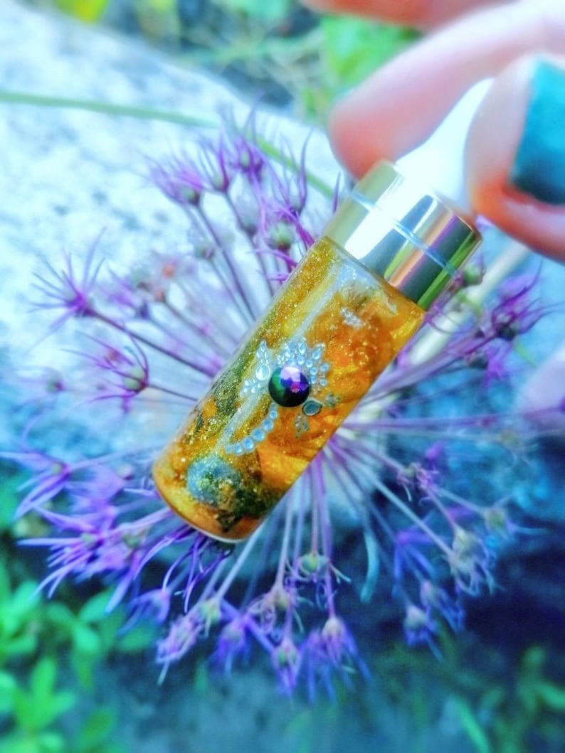 Op de afbeelding: Een klein, doorzichtig glazen flesje met een gouden dop en een druppelaar. Het flesje is gevuld met een gouden vloeistof en bevat glitter en een kleine, iriserende edelsteen. Het flesje ligt op een paarse bloem.