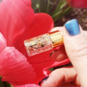 May include: A small clear glass bottle with a gold lid and gold glitter inside. The bottle is being held by a hand with blue nail polish.
