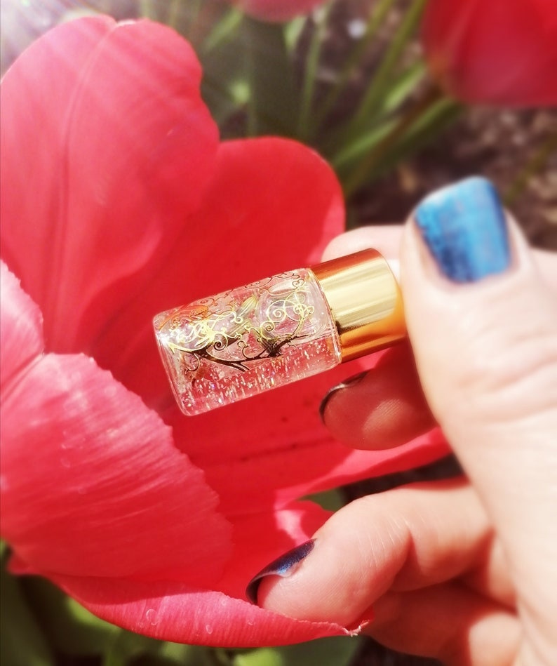 May include: A small clear glass bottle with a gold lid and gold glitter inside. The bottle is being held by a hand with blue nail polish.