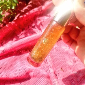 Può includere: Un tubetto di lucidalabbra trasparente riempito con un liquido scintillante, arancione dorato. Il tubetto ha un tappo argentato e un piccolo emblema decorativo. Il lucidalabbra è appoggiato su un tessuto di pizzo rosa e un panno rosso.