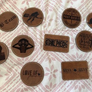 May include: A collection of ten brown leather patches with stitched edges, featuring various laser-engraved designs including text like B Team, One Piece, Silva Attorneys, and religious quotes, alongside illustrations of characters and symbols.