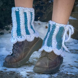 Puede incluir: Calentadores de piernas de ganchillo a rayas turquesa y blancas, usados sobre botas de cuero marrón. Los calentadores tienen un borde festoneado y están atados con hilo blanco. Las botas están atadas.